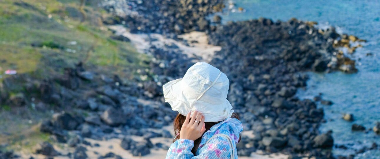 Woman in hat sitting on cliff with view of rocky coast, wearing floral jacket, capturing tranquility.